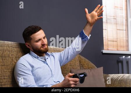 A man at home sitting on the couch with a joystick in his hands. The disgruntled man is outraged about his loss. Spending time. Stock Photo