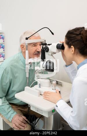 Young brunette female ophthalmologist in whitecoat testing eyesight of senior male patient while using medical equipment in clinics Stock Photo