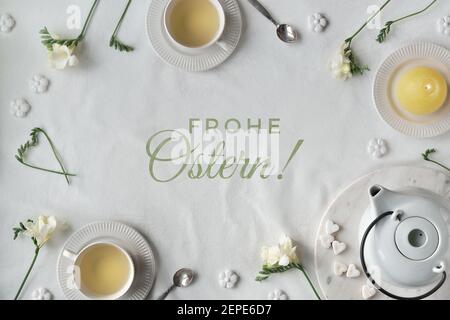 Text Frohe Ostern means Happy Easter in English. Springtime background. Spring freesia flowers, tea cups and teapot. Easter eggs and sugar hearts. Fla Stock Photo