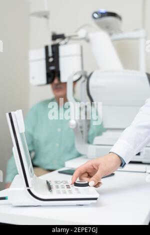 Hand of young contemporary ophthalmologist turning knob on examination equipment while checking eyesight of senior man Stock Photo