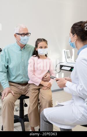 Senior man and his granddaughter in protective masks consulting with young female professional ophthalmologist in contemporary clinics Stock Photo