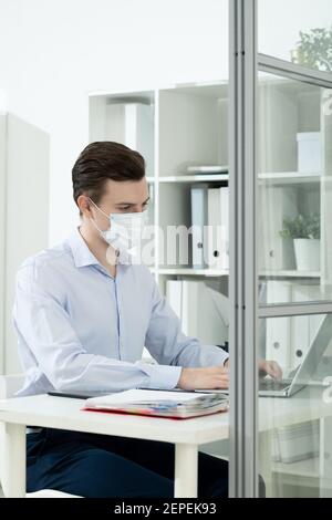 Young businessman in protective mask and formalwear looking at laptop display while working with data in office against shelves with folders Stock Photo