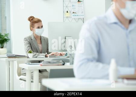 Young elegant businesswoman or creative designer in protective mask looking at computer screen in office in front of her male colleague Stock Photo