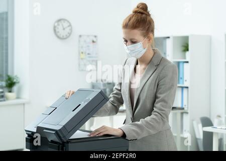 Young secretary in grey suit and protective mask putting paper in xerox machine to make copies against shelves with documents in office Stock Photo