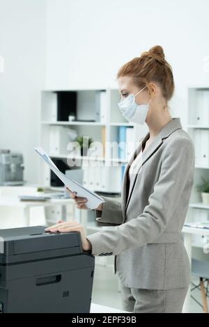Young secretary in grey suit and protective mask looking through one of papers while standing by xerox machine in office environment Stock Photo