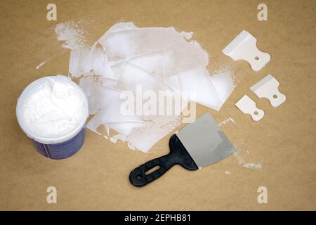 A bucket of white putty and a spatula on a fiberboard board top view. Putty strokes to create a background for a photo. Rubber and metal spatulas for Stock Photo