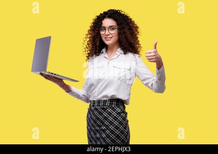 Business woman holds laptop and shows thumb up. Female in white shirt on yellow isolated background Stock Photo