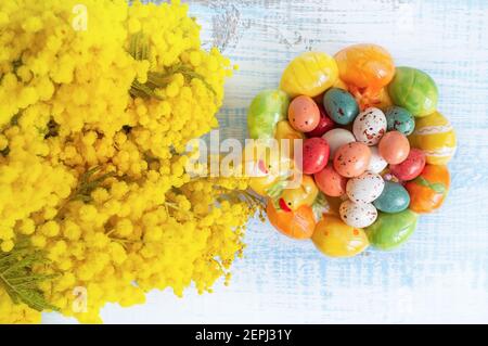 Easter composition. Candy in the shape of Easter eggs on a beautiful ceramic Easter plate. Next to the plate is a bouquet of yellow mimosa. Congratula Stock Photo
