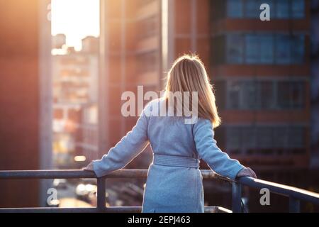 Businesswoman standing on terrace and looking at skyscraper in city. Blond hair woman resting at balcony in modern city at Europe. Fashionable lady Stock Photo
