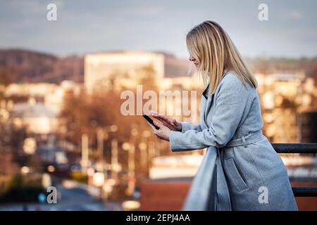 Businesswoman using her smartphone in city. Woman standing at terrace and texting message on mobile phone. Leadership and communication by phone Stock Photo
