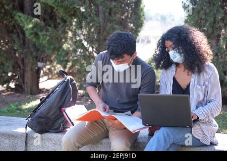 Two latin students wearing protective face mask studying together sitting on a bench outdoor. New normal at university campus. Stock Photo