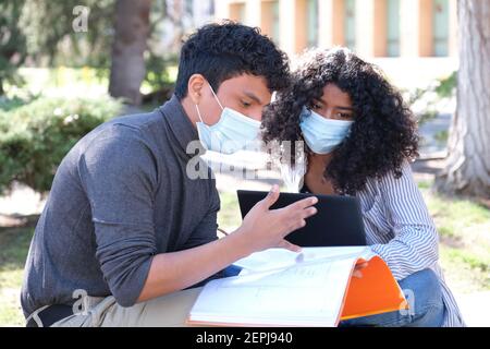 Two latin students wearing protective face mask studying together sitting on a bench outdoor. New normal at university campus. Stock Photo
