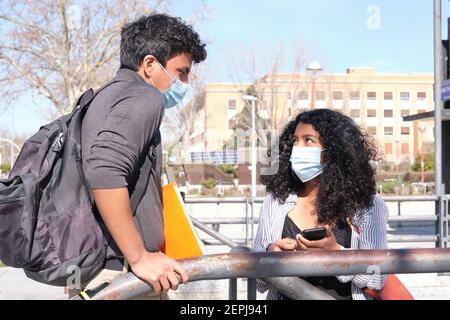 Young latin couple wearing protective face mask looking each other and talking at a train station. New normal in the university campus. Stock Photo