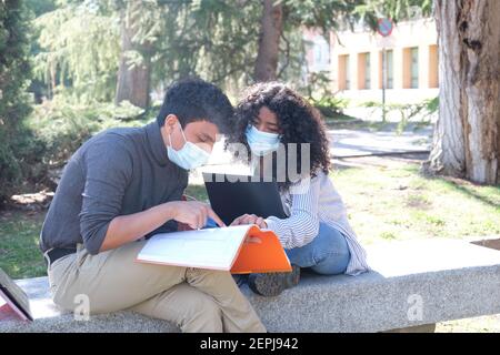 Two latin students wearing protective face mask studying together sitting on a bench outdoor. New normal at university campus. Stock Photo