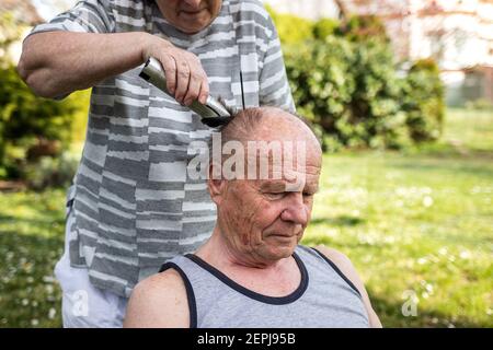 Woman is doing haircut with hair clipper to senior man outdoors at garden. Elderly people take care each other. Senior couple Stock Photo