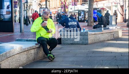A man in a fluorescent jacket sits alone on a marble bench, drinks a takeaway drink and checks his mobile phone in Broad Street, Reading, Stock Photo