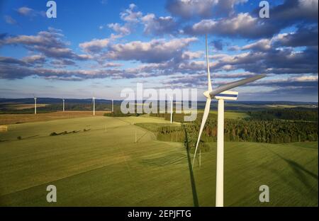 Aerial view of wind turbine farm. Wind power plants in green landscape against sunset sky with clouds. Aerial, drone inspection of wind turbine. Stock Photo