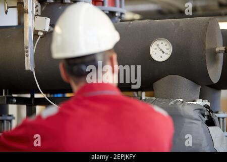 Energy industry. A technician dressed in red overalls and a white helmet checking the heating parameters. Heating water distribution technology.. Stock Photo