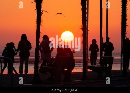 Southend on Sea, Essex, UK. 27th Feb, 2021. The warm sunny day has ended with a glowing sunset behind the palm trees and visitors along the seafront at Southend on Sea. People walking and sitting on the promenade of Western Esplanade as the sun goes down during the COVID 19 lockdown Stock Photo