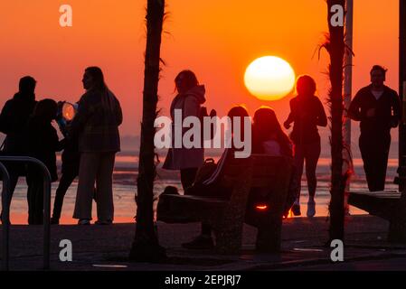 Southend on Sea, Essex, UK. 27th Feb, 2021. The warm sunny day has ended with a glowing sunset behind the palm trees and visitors along the seafront at Southend on Sea. People walking and sitting on the promenade of Western Esplanade as the sun goes down during the COVID 19 lockdown. Evening run Stock Photo