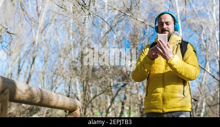 young hipster man with a beard and backpack on his back listening to music with his headphones and his mobile phone in a park Stock Photo