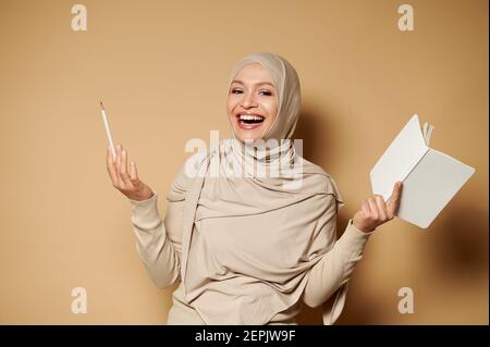 Muslim woman in hijab holding a notepad and pencil and laughing while posing to camera on beige background with copy space Stock Photo