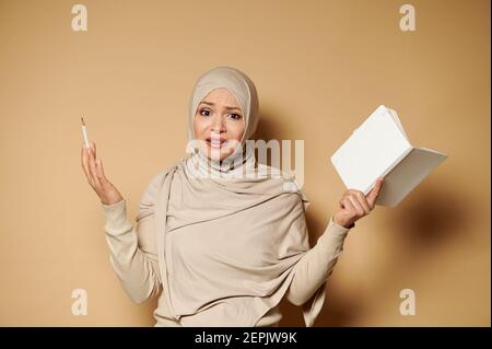 Muslim woman expressing surprise, dissatisfactrion standing against beige background with notepad and pencil in hands Stock Photo