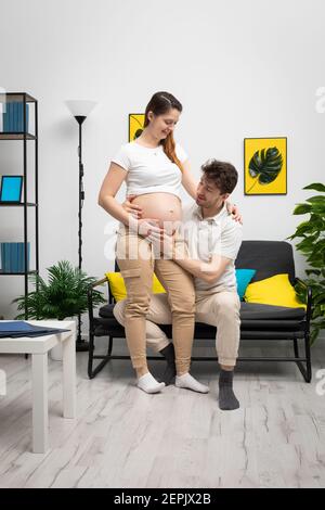 At home, a young married couple watch the unborn child move in the pregnant woman's belly. Living room decorated in a modern style with bright lighting. Stock Photo