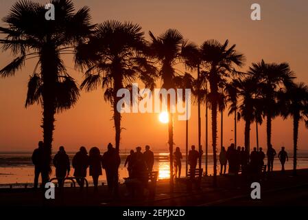 Southend on Sea, Essex, UK. 27th Feb, 2021. The warm sunny weather has attracted visitors to Southend, with the seafront roads especially busy. People are walking the seafront promenades into the evening as the sun set behind the palm trees. People walking and sitting on the promenade of Western Esplanade as the sun goes down during the COVID 19 lockdown Stock Photo