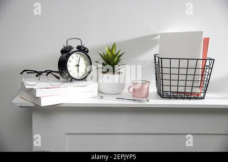 A stack of books, an artificial succulent plant in a ceramic pot, glasses, a metal basket with notebooks, a pen and a cup on a white table surface. Co Stock Photo