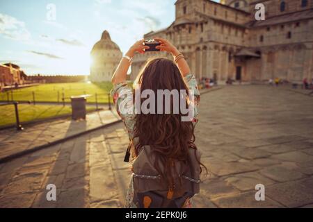 Seen from behind young woman in floral dress with vintage camera and backpack near Duomo di Pisa. Stock Photo
