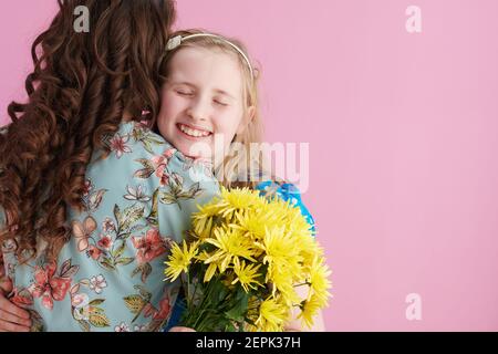 smiling elegant mother and daughter with long wavy hair with yellow chrysanthemums flowers embracing against pink background. Stock Photo