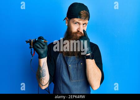 Redhead man with long beard tattoo artist wearing professional uniform and gloves pointing to the eye watching you gesture, suspicious expression Stock Photo
