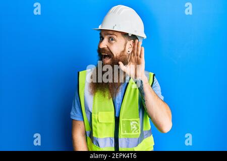 Redhead man with long beard wearing safety helmet and reflective jacket smiling with hand over ear listening an hearing to rumor or gossip. deafness c Stock Photo