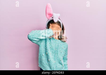 Little beautiful girl wearing cute easter bunny ears smiling and laughing with hand on face covering eyes for surprise. blind concept. Stock Photo