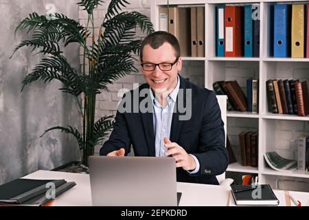 Young cheerful man businessman with glasses using laptop sitting in office at the table wearing suit, he is smiling looking into laptop Stock Photo