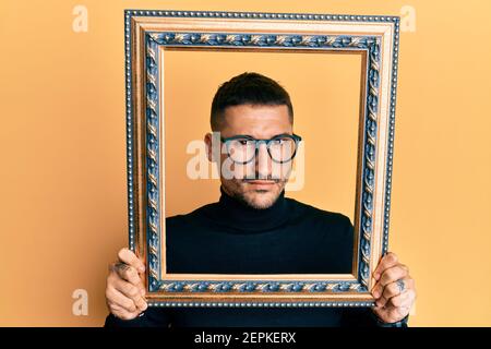 Handsome man with tattoos holding empty frame skeptic and nervous, frowning upset because of problem. negative person. Stock Photo