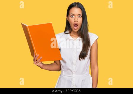 Beautiful hispanic woman reading book scared and amazed with open mouth for surprise, disbelief face Stock Photo