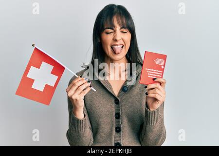 Young hispanic woman holding swiss flag and passport sticking tongue out happy with funny expression. Stock Photo