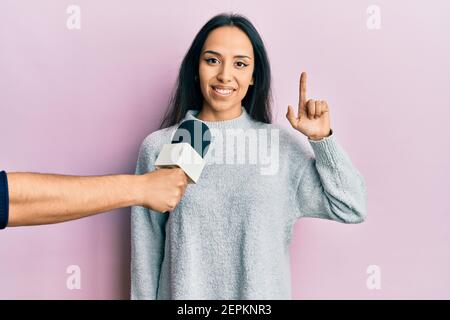 Young hispanic girl being interviewed by reporter holding microphone smiling with an idea or question pointing finger up with happy face, number one Stock Photo