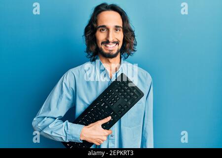 Young hispanic man holding keyboard looking positive and happy standing and smiling with a confident smile showing teeth Stock Photo