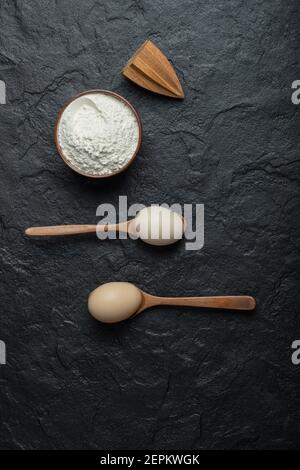 Spoons of eggs and bowl of flour on dark background Stock Photo