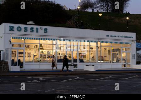 Rossi's ice cream parlour on Western Esplanade, Southend on Sea, Essex, UK, at dusk with lights on inside. People passing and sitting on top of cliffs Stock Photo