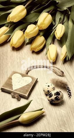 Easter zero waste arrangement with bunch of yellow tulips, wooden heart and quail eggs on wooden table. This image is toned. Stock Photo