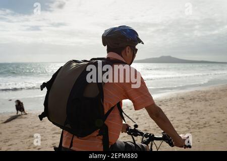 A cyclist riding along Milford beach with Rangitoto Island in the background and a dog playing on the beach