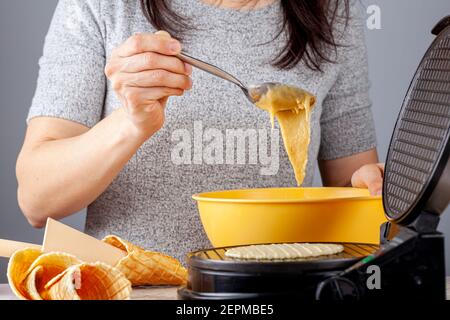 A woman is  using a non stick electrical waffle cone maker to make homemade ice cream cones. Fresh made hand rolled cones are lying on marble countert Stock Photo