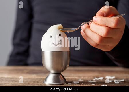 A small child is eating soft boiled humpty dumpty egg placed in egg cup on a wooden breakfast table. She uses spoon to eat the egg from its shell. She Stock Photo