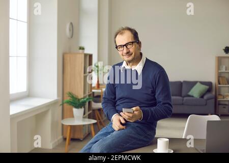 Happy adult businessman using mobile phone sitting on edge of desk in his home office Stock Photo