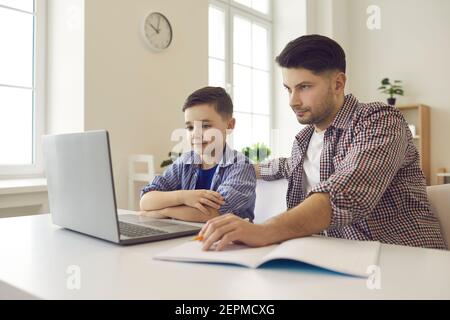 Dad sits with his son and helps him with his studies using a laptop to study school lessons. Stock Photo