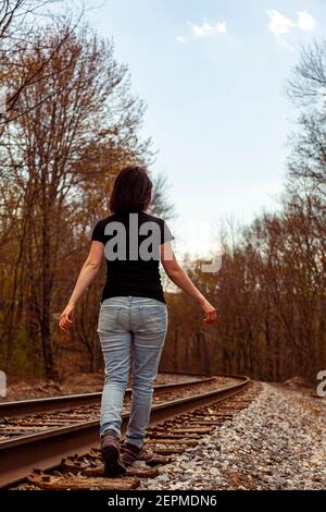A young caucasian woman wearing jeans and t shirt is walking on the railroad tracks at a rural location with her arms wide open. An abstract moody con Stock Photo